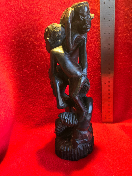 A sculpture of an African figure seated with a child on its lap, positioned against a red background with a measuring tape in the background.